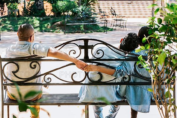 Rear view of unfaithful woman holding hands with another man while boyfriend hugs her. Unfaithful couple holding hands on a park bench. Cheating girlfriend with her boyfriend holding another man hand