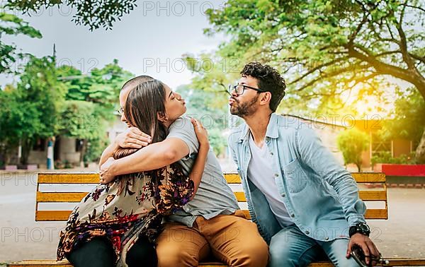 Unfaithful woman sitting hugging her boyfriend and secretly kissing another man. Unfaithful girlfriend hugging her boyfriend in a park secretly kissing another man