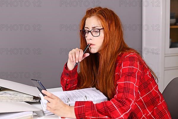 Woman with red hair sitting at desk with document ring files looking at mobile phone in hand