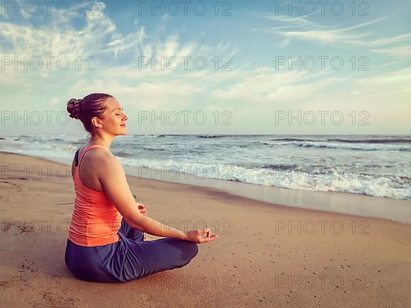 Vintage retro hipster effect image of woman doing yoga meditating and relaxing in Padmasana Lotus Pose with chin mudra outdoors at tropical beach on sunset