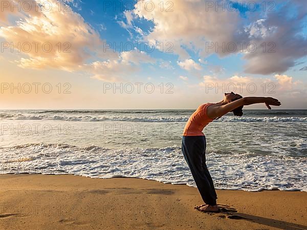 Young sporty fit woman doing yoga Sun salutation Surya Namaskar pose Hasta Uttanasana on tropical beach on sunset