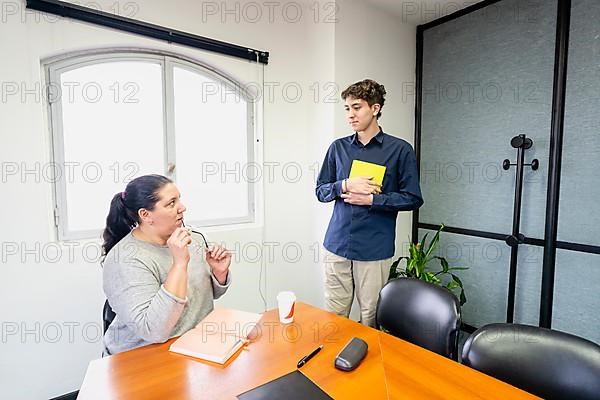 Businesswoman sitting at the head of the meeting table looking at her assistant standing next to her