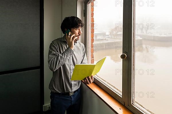 Latino businessman working in an office overlooking the river. Talking on the phone and reviewing notes while he looks out the window