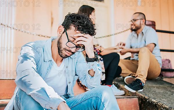 Sad boyfriend sitting on some stairs while his girlfriend talking to another man. Sad man seeing his girlfriend cheating with his friend. Girl talking to another man and her boyfriend is offended