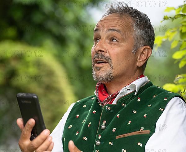 Man in Bavarian traditional traditional costume with mobile phone in hand looks astonished