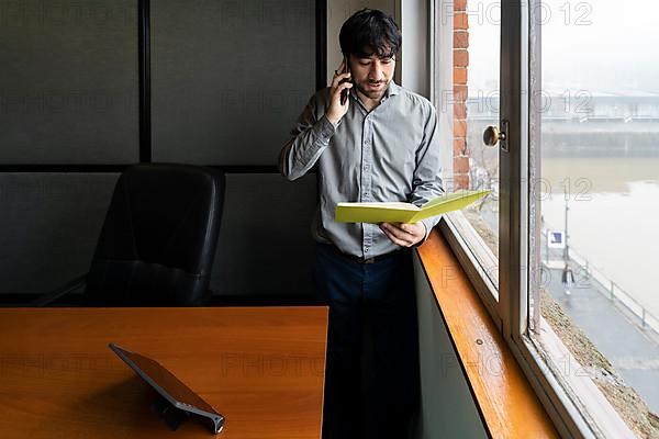 Latino businessman working in an office overlooking the river. Talking on the phone and reviewing notes