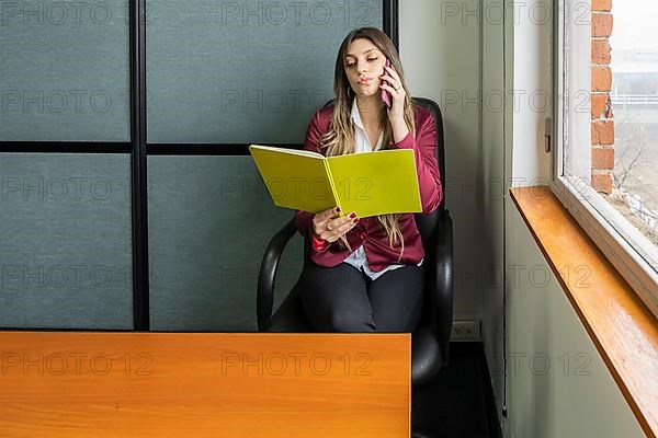 Young blonde business woman sitting in her office using the phone while checking notes