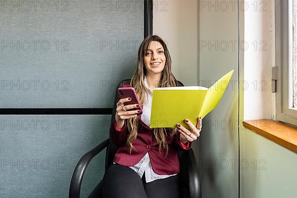 Young blonde business woman sitting in her office using the phone while checking notes looking at the camera