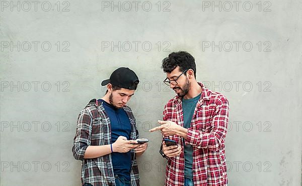 Two young friends leaning against a wall checking their cell phones