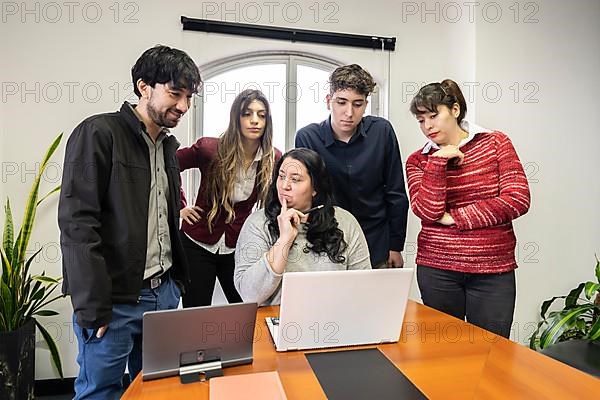 Group of employees standing around their boss at the meeting table looking at her notebook screen