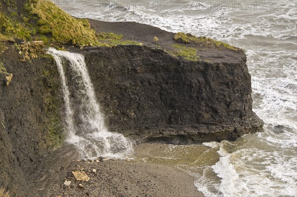 Waterfall flowing over hard shale to beach on rough sea coast