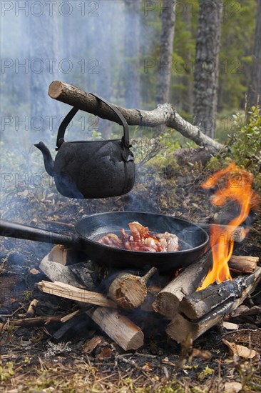 Blackened tin kettle boiling water and pan cooking bacon over flames from campfire