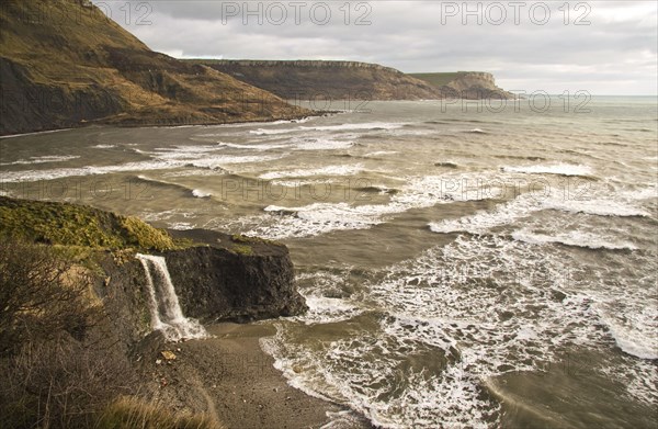 View of coastline with waterfall flowing over hard shale to beach on coast with rough sea