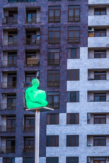 Illuminated human sculpture on a pole in front of a building seen from the Yerevan Cascade