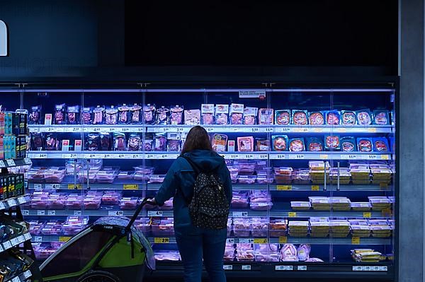 A woman with a pram is standing in front of the meat and sausage cooling shelf