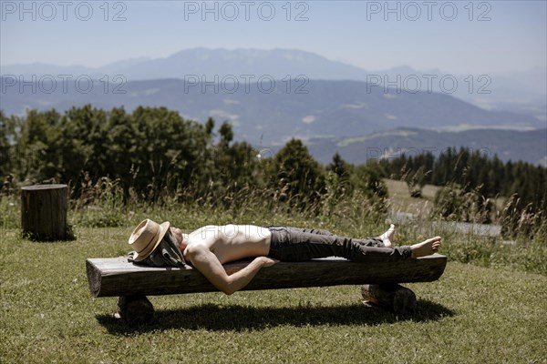 Man with hat and naked upper body lying on a wooden bench on the mountain pasture