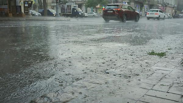 Raindrops fall on the road with cars driving in the background. Heavy torrential rain in the city