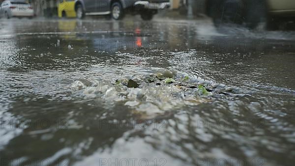 A stream of rainwater flows into the storm drain cars are driving in the background. Heavy torrential rain in the city