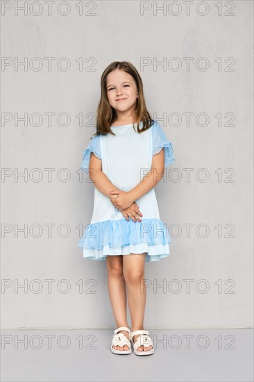 Cute little girl in blue dress and white sandals posing with hands clasped down near the gray wall