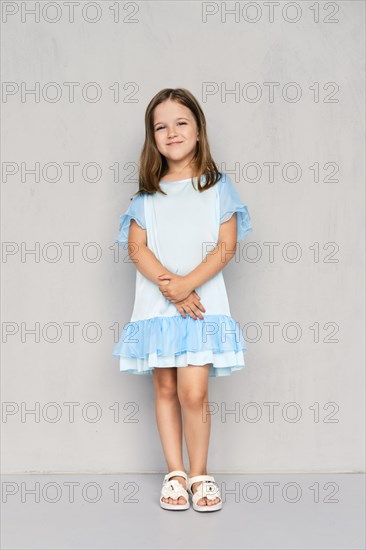 Cute little girl in blue dress and white sandals posing with hands clasped down near the gray wall