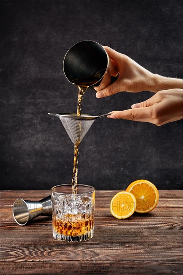 Hand of bartender with shaker and strainer pouring cocktail with brandy and orange bitter