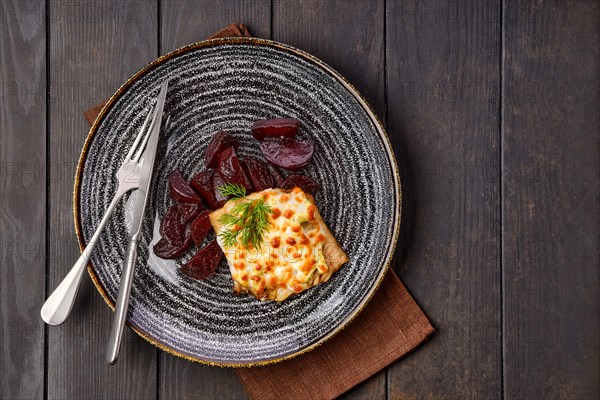 Top view of fried cod fillet with melted cheese topping and roasted beetroot slices on dark wooden table