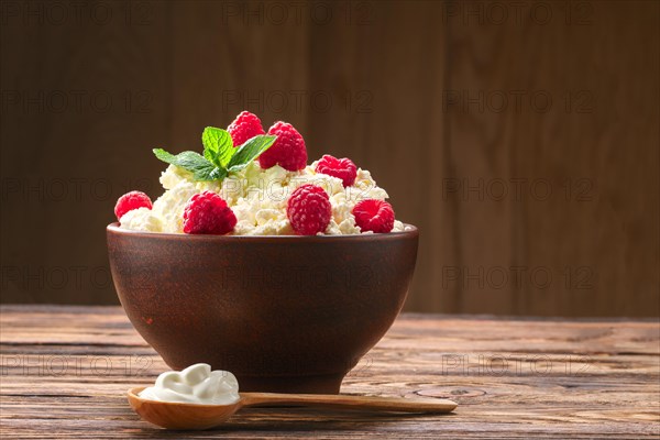 Wooden spoon with sour cream near clay plate with fresh cottage cheese with raspberries and mint