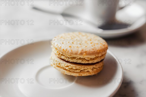 Close up photo of macaron with salted caramel on a white saucer with blurred cup of coffee on the background