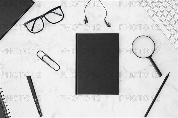 Closed black diary with eyeglasses earphone magnifying glasses pencil keyboard diary white desk