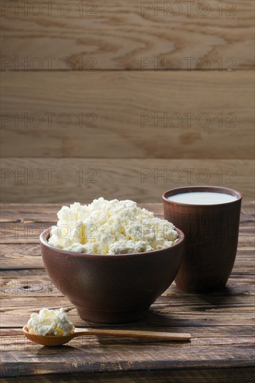 Fresh cottage cheese in clay bowl with wooden spoon with a glass of milk on rustic wooden background