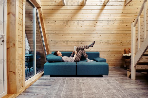 Middle aged man relaxing with book on sofa at chalet. Man in checkered trousers