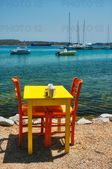 Yellow cafe restaurant table of street cafe with chairs on beach in Adamantas town on Milos island with Aegean sea with boats and yachts in background. Milos island