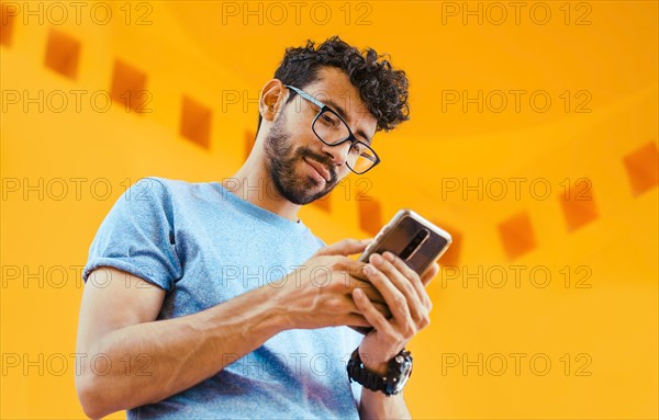 Low angle view of young man using cell phone outdoors. Guy sending a message with a cell phone near a yellow wall outdoors. Lifestyle of handsome bearded man using cell phone outdoors