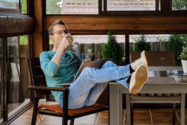 Middle aged man drinks wine while watching online broadcast on laptop sitting on the veranda of his country house