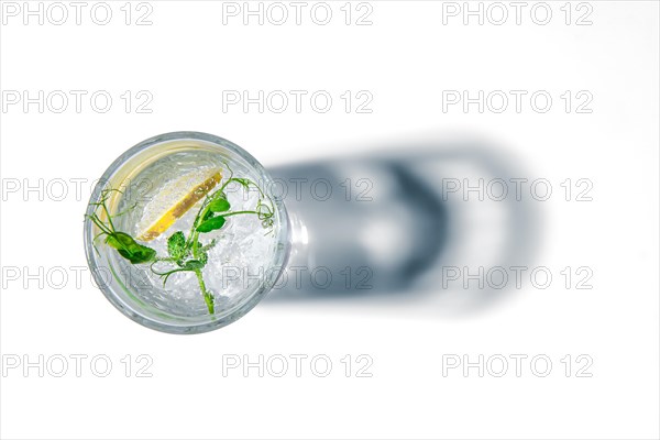 Overhead view of glass with cold water with ice and lemon casting a shadow on white background