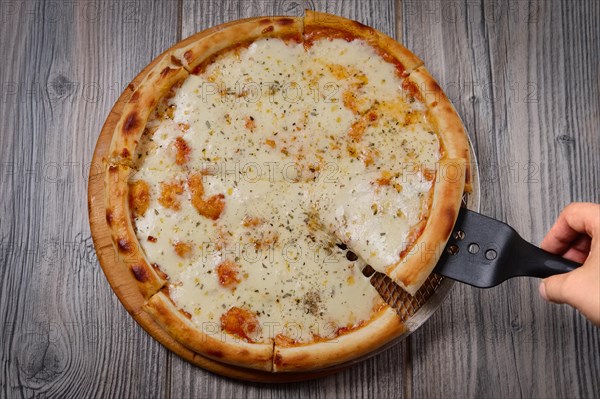 Top view of hand with spatula taking up slice of pizza with cheese on wooden table