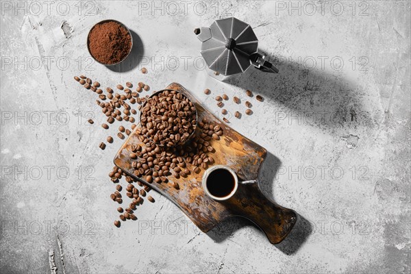 Overhead view of cup of espresso with coffee beans scattered on wooden board and a pot under hard morning sun