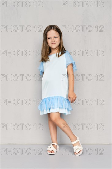 Cute little girl in blue dress and white sandals posing near the gray wall