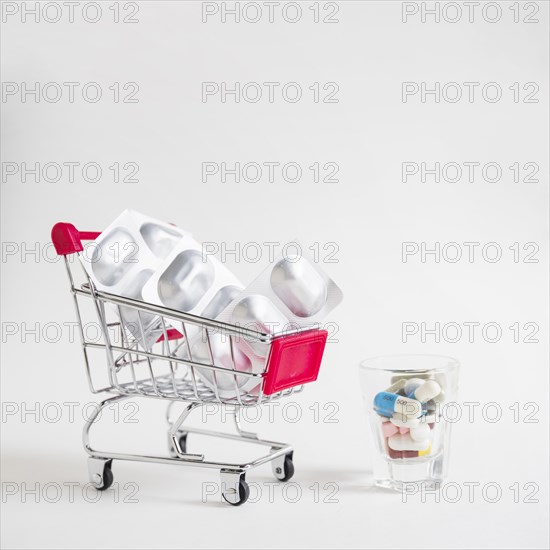 Many pills small glass with shopping cart with silver blister pills white background