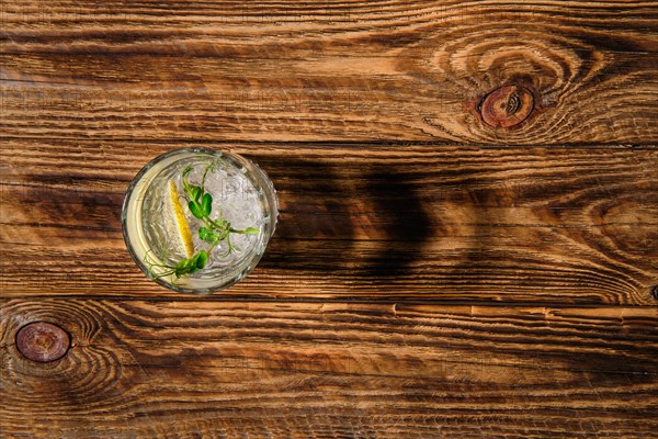 Overhead view of glass with cold water with ice and lemon casting a shadow on wooden table