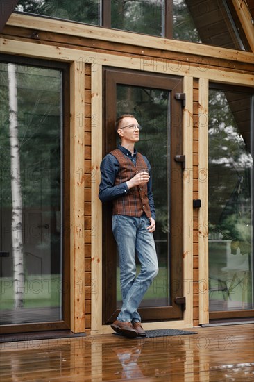 Pensive middle age man stands on the terrace of a wooden bungalow and drinks bourbon