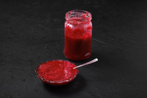 Jar and saucer with cherry jam on black background