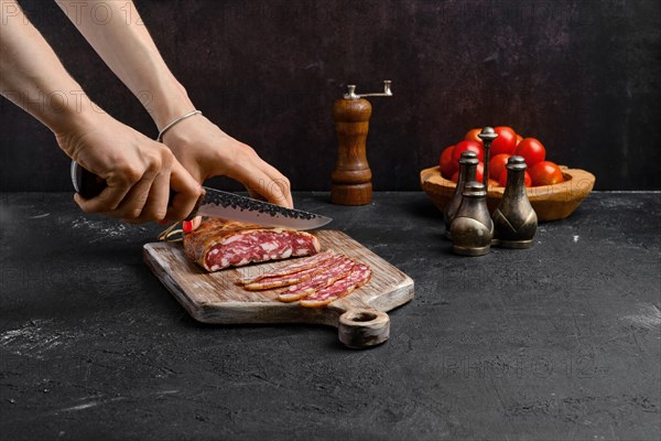 Female hand with knife slicing smoked pork sausage in organic casing on wooden cutting board