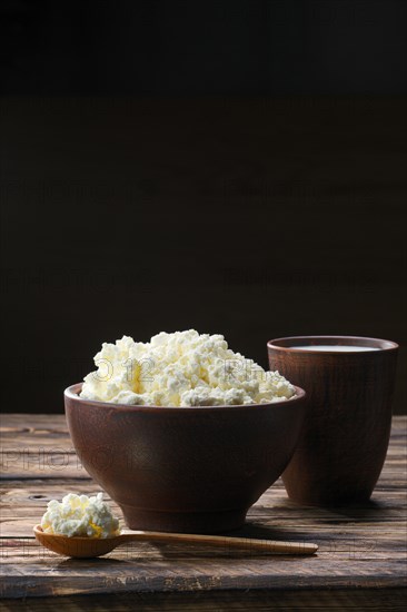 Fresh cottage cheese in clay bowl with wooden spoon with a glass of milk on rustic wooden background