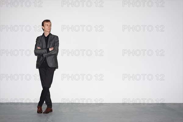 Middle age man in gray wool jacket and brown shoes standing near white concrete wall with crossed hands