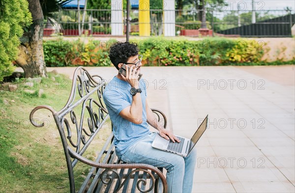 Young man sitting in a park working with his laptop and cell phone. Man in a park working online with laptop and calling on cellphone. Freelancer man working with laptop while calling on the phone