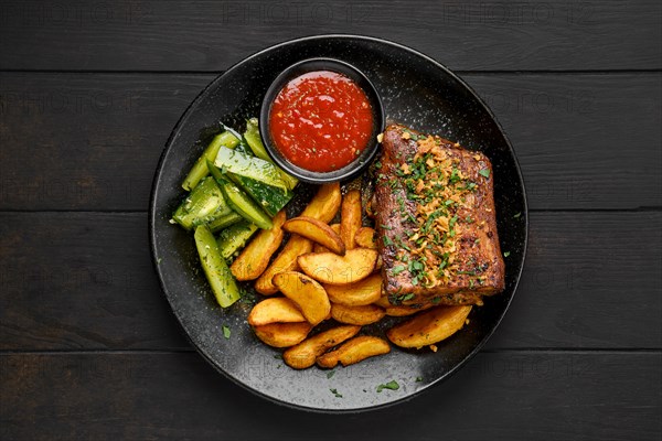 Top view of roasted beef ribs with potato wedges and pickled cucumber on a plate