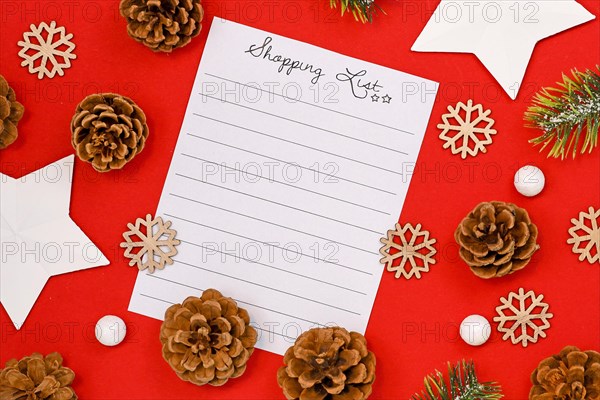 Concept for Christmas shopping with top view flat lay with empty shopping check list and seasonal decorations like pine cones and stars on bright red background