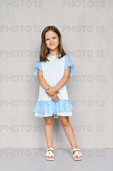 Cute little girl in blue dress and white sandals posing with hands clasped down near the gray wall