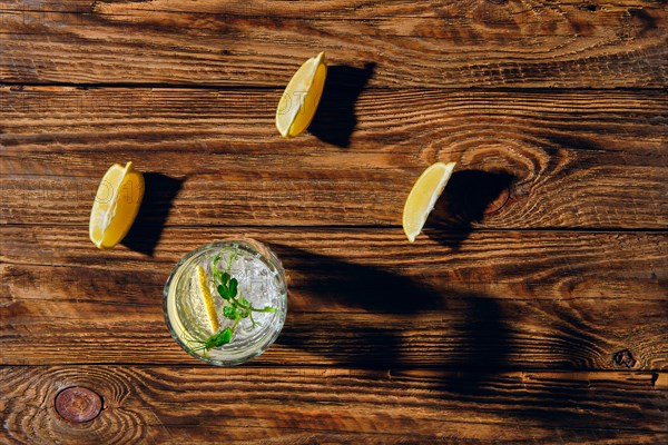 Overhead view of glass with cold water with ice and lemon casting a shadow on wooden table